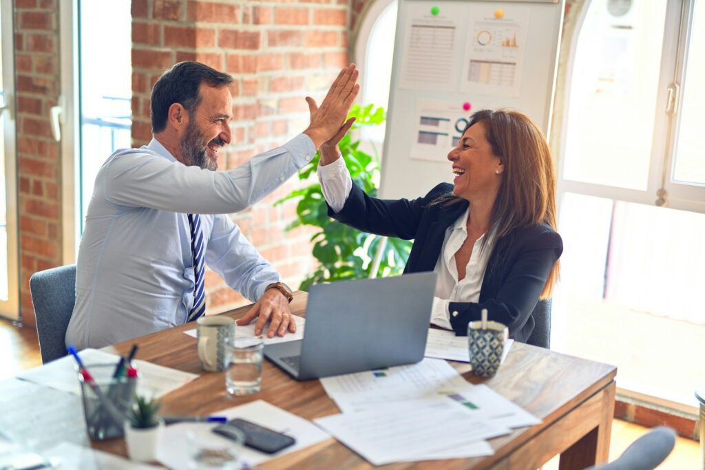 Two people in a workplace hi-fiving each other over a workdesk and a laptop